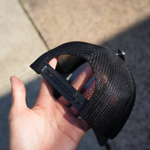 Black snap back flex fit mesh cap held by a hand on a concrete surface