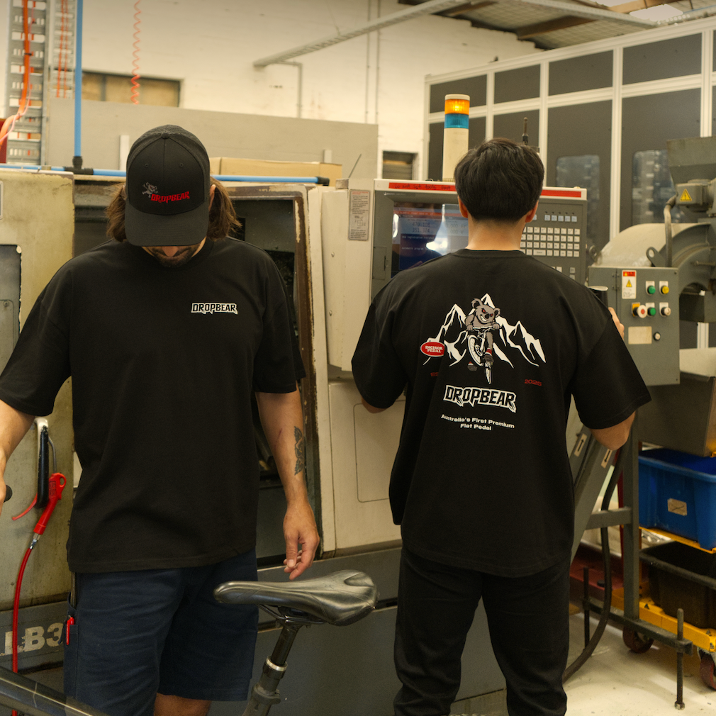 Two people in a workshop setting with machinery and equipment. They are wearing Dropbear Oversized Tees
